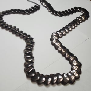 May include: A silver-toned necklace with a series of connected, heart-shaped links. The necklace is laid out on a white surface, with the clasp open. The links have a reflective, metallic finish.