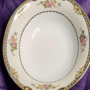 May include: Oval, off-white porcelain serving dish with gold handles and trim. The rim features a floral and scroll design in pink, yellow, blue, and gold. The center of the dish is plain white.