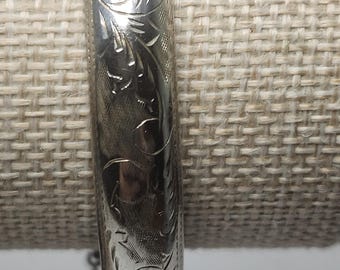 May include: A silver-toned metal bangle bracelet featuring an intricate engraved floral pattern across its curved surface. The bracelet has a polished finish and includes a thin, delicate safety chain attached to one side of the hinged closure mechanism.
