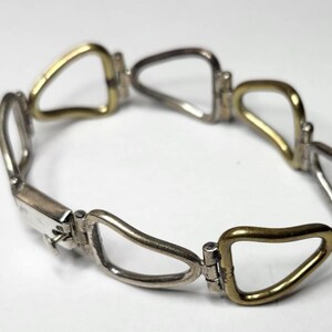 May include: A silver and gold-toned bracelet with a unique linked design. The bracelet features a series of open, teardrop-shaped links, alternating between silver and gold-colored metal. The clasp is a rectangular silver piece.