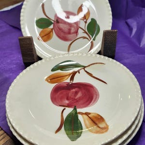 May include: A stack of off-white ceramic plates with a red apple and green and orange leaves design. The plates have a beaded edge. Two plates are displayed on a wooden stand, with a purple background.