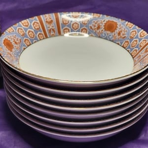 May include: A stack of white ceramic bowls with a decorative rim featuring orange and blue floral patterns. The bowls have a gold trim and are stacked on top of each other. The background is a solid purple color.