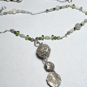 Könnte beinhalten: Eine silberne Halskette mit einer Perlenkette und einem Anhänger. Die Kette hat weiße, grüne und schwarze Perlen. Der Anhänger hat einen strukturierten silbernen Ball und drei silberne, muschelförmige Anhänger mit einer Abalone-Muschel am Boden.