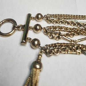 May include: Gold-toned multi-strand necklace with a toggle clasp and a decorative tassel. The necklace features multiple chains and beaded accents. The tassel is a light gold color.