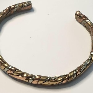 May include: A mixed-metal cuff bracelet with a twisted rope design. The bracelet features copper, silver, and gold-toned metal strands woven together. The open-ended design allows for easy wear. The bracelet is shown on a white background.