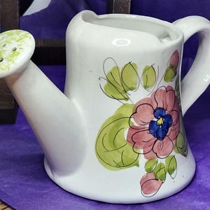 May include: White ceramic watering can with a green-spotted spout and a curved handle. The can is decorated with pink and blue floral designs and green leaves. A charming garden-themed decorative piece.