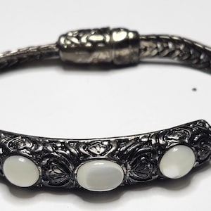 May include: A silver-toned bracelet with a braided band and a decorative centerpiece. The centerpiece features three oval, white cabochon stones set within an ornate, floral-inspired design. The bracelet has a secure clasp.