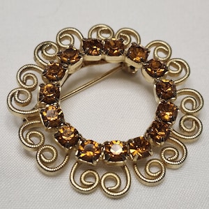 May include: A vintage gold-toned brooch with a circular design. The center features a ring of amber-colored rhinestones. The outer edge has decorative swirls. A classic piece of jewelry.
