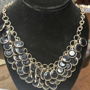 May include: A gold-toned chain necklace with multiple dark blue, circular pendants. The pendants are arranged in a cascading pattern, creating a layered effect. The necklace is displayed on a black velvet bust.