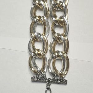 May include: A silver and gold-tone chain bracelet. The bracelet features two rows of interlocking oval links in alternating silver and gold colors. The clasp and extender chain are silver.