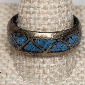 May include: A silver-toned ring with a geometric design. The ring features a band inlaid with small, bright blue stones, creating a striking contrast against the silver metal. The ring is displayed on a textured, off-white surface.