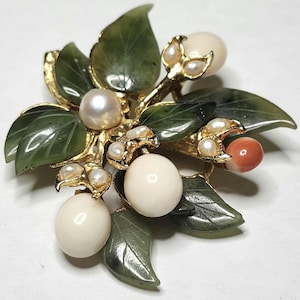 May include: A vintage brooch featuring a floral design with green jade leaves, ivory-colored orbs, small pearls, and a coral-colored orb. The brooch has a gold-tone metal frame.