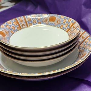 May include: A stack of white ceramic bowls with a decorative rim featuring a blue, orange, and gold pattern. The bowls are stacked on a matching plate, creating a cohesive set. The set is on a purple surface.