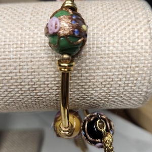 May include: Gold-toned bracelet with decorative glass beads. The beads feature intricate designs in green, pink, and gold. A tassel hangs from one of the beads, adding a touch of elegance. The bracelet is displayed on a neutral-toned surface.