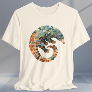 May include: A white t-shirt with a colorful graphic of a curled up pangolin. The pangolin is made up of geometric shapes and has a blue, green, orange, and brown color scheme.
