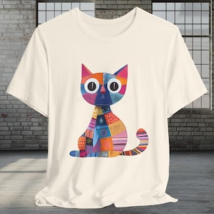May include: Cream-colored t-shirt featuring a colorful patchwork cat design. The cat has large, round eyes and a variety of patterned patches in shades of blue, orange, pink, and green. The shirt is made of a soft material.