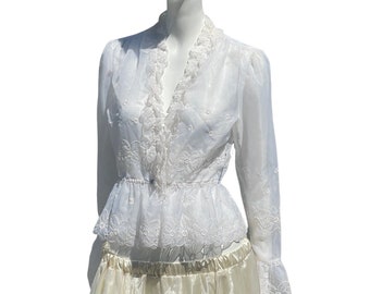 May include: A white sheer blouse with a lace trim and embroidered floral details. The blouse has a V-neckline and long sleeves with ruffled cuffs. It is paired with a cream-colored skirt.