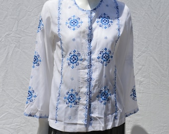 Puede incluir: Blusa blanca con patrones florales bordados en azul. La blusa tiene un frente abotonado y mangas largas.