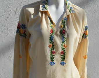 May include: A cream-colored blouse with long sleeves and a V-neck. The blouse is embroidered with colorful floral designs in shades of red, blue, green, and yellow.