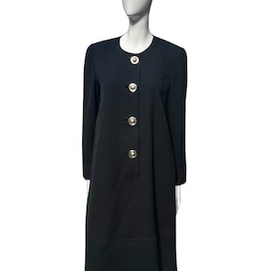 Op de afbeelding: Een zwarte jurk met lange mouwen en een ronde halslijn. De jurk heeft grote, zilverkleurige knopen aan de voorkant en decoratieve stiksels bij de manchetten en zoom. De jurk wordt op een mannequin gepresenteerd.