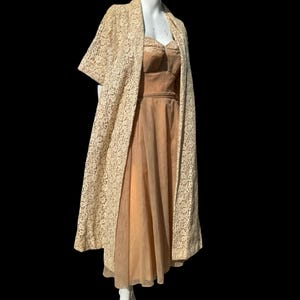 May include: A vintage lace dress with a long, flowing skirt and a fitted bodice. The dress is a pale cream color and has a delicate floral pattern. It is paired with a matching lace jacket with short sleeves.