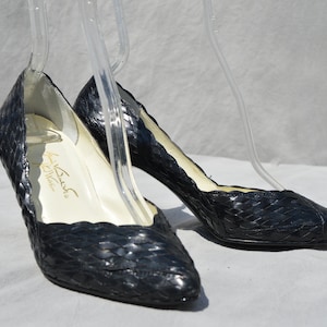 Pu&ograve; includere: Un paio di scarpe con tacco in pelle nera intrecciata con punta a punta. Le scarpe sono su una superficie bianca e hanno un'etichetta dorata all'interno che recita "La  Bush  California".
