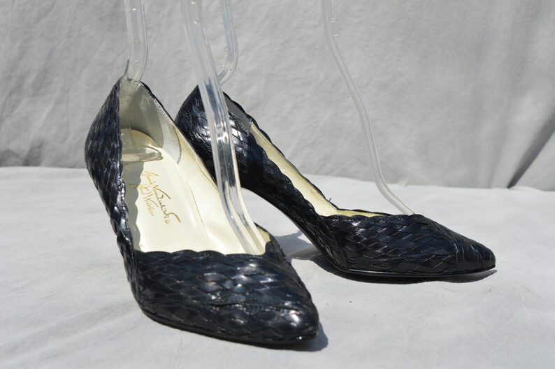 Pu&ograve; includere: Un paio di scarpe con tacco in pelle nera intrecciata con punta a punta. Le scarpe sono su una superficie bianca e hanno un'etichetta dorata all'interno che recita "La  Bush  California".