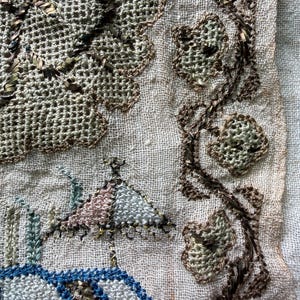 May include: Close-up of a vintage textile with intricate embroidery. The design features a landscape with a building and a tree, along with floral elements. The embroidery uses shades of blue, pink, green, and brown on a beige background.