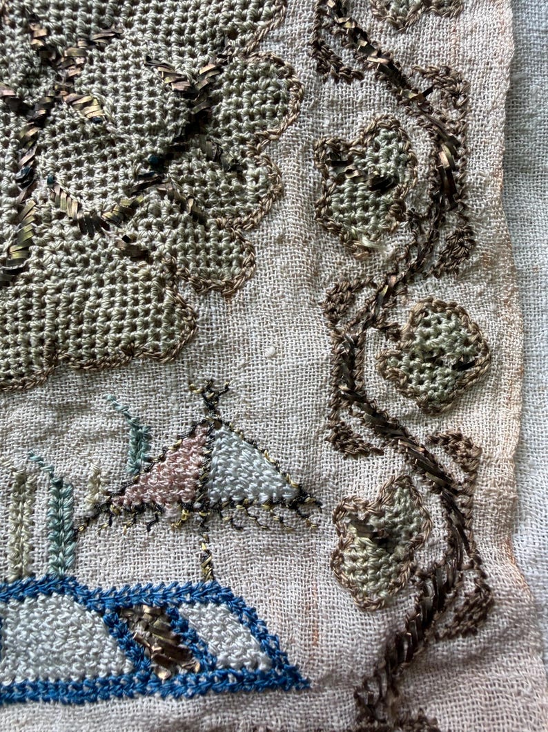 May include: Close-up of a vintage textile with intricate embroidery. The design features a landscape with a building and a tree, along with floral elements. The embroidery uses shades of blue, pink, green, and brown on a beige background.