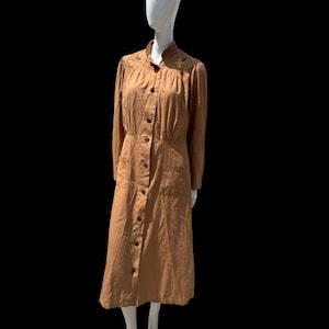 May include: A tan-colored vintage dress with long sleeves and a button-down front. The dress features embroidered floral details on the collar and shoulders. The dress is displayed on a mannequin.