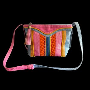 May include: A colorful shoulder bag with a pink strap and a matching pink tassel. The bag features a patchwork design with vertical stripes in pink, orange, yellow, and blue. The bag is set against a black background.