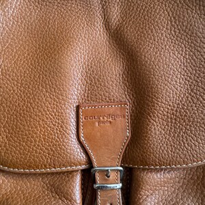 Puede incluir: Una mochila de cuero marr&oacute;n con una hebilla plateada y un parche cosido blanco que dice "Courr&egrave;ges Paris".
