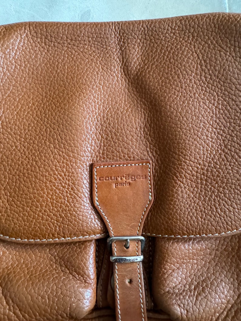 Puede incluir: Una mochila de cuero marr&oacute;n con una hebilla plateada y un parche cosido blanco que dice "Courr&egrave;ges Paris".