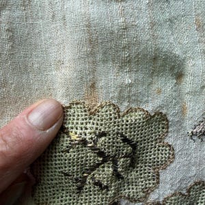 May include: Close-up of a section of fabric with embroidered floral designs. The embroidery features a light green color with brown accents, creating a textured appearance. The fabric has a light beige color with some brown spots.