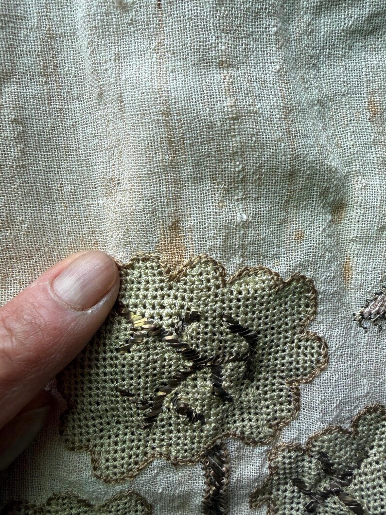 May include: Close-up of a section of fabric with embroidered floral designs. The embroidery features a light green color with brown accents, creating a textured appearance. The fabric has a light beige color with some brown spots.