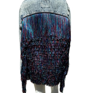 Puede incluir: Una chaqueta vaquera de color azul claro con un ribete de flecos. Los flecos están hechos de hilo multicolor en tonos azules, rosas y morados.