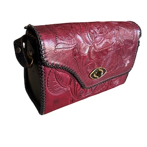 Könnte beinhalten: Burgunderfarbene Leder-Umhängetasche mit geprägtem Blumenmuster. Die Tasche hat einen schwarz genähten Besatz, einen goldfarbenen Verschluss und einen schwarzen Lederriemen. Die Tasche hat eine rechteckige Form.