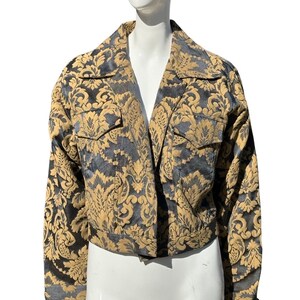 May include: A cropped jacket featuring a gold and black damask pattern. The jacket has a collar, long sleeves, and a button fastening.