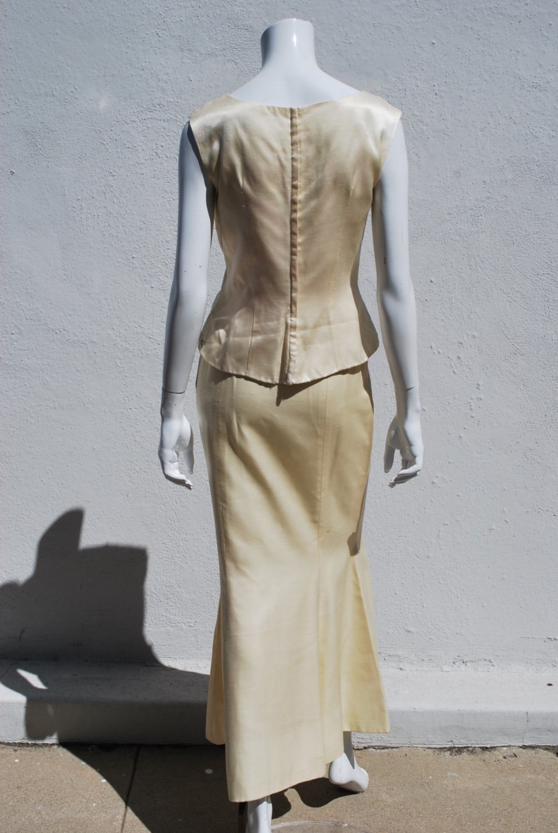 Op de afbeelding: Een mouwloze, ivoorwitte satijnen jurk met een getailleerd lijfje en een zwierige rok. De jurk heeft een rits aan de achterkant en een klein peplum detail op de taille.