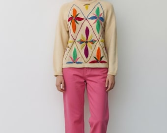 May include: A woman wearing a cream-colored sweater with a colorful floral design and pink pants. The sweater features a diamond pattern with embroidered flowers in orange, green, blue, and purple. The pants are a vibrant pink, and the shoes are pink flats.