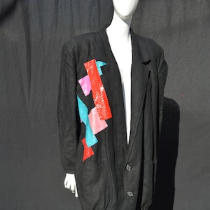 Może przedstawiać: Czarny blazer z geometrycznym wzorem z czerwonymi, różowymi i turkusowymi akcentami. Blazer ma długi, luźny krój i zapięcie na jeden guzik.