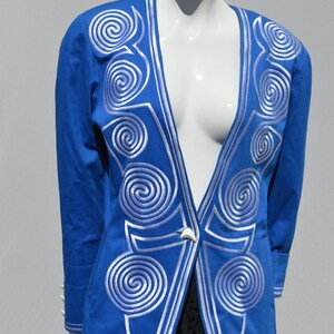 Puede incluir: Una chaqueta azul con bordados en espiral blancos en la parte delantera y las mangas. La chaqueta tiene un cuello en V profundo y un cierre de un solo botón.