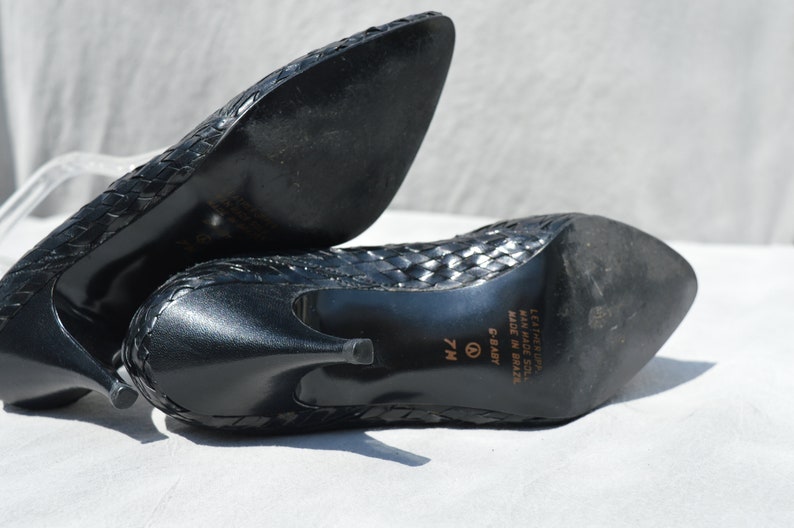 Pu&ograve; includere: Scarpe con tacco in pelle nera intrecciata con punta a punta. Le scarpe hanno un'etichetta di misura 7M sulla suola e riportano "Leather Upper, Man Made Sole, Made in Brazil, C-Balt" sul fondo.