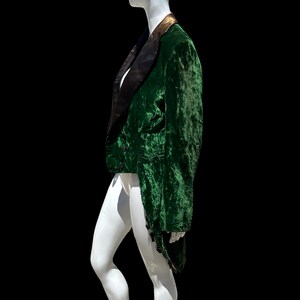 K&ouml;nnte beinhalten: Eine gr&uuml;ne Samtjacke mit schwarzem Satinrevers und goldenen Verzierungen. Die Jacke hat einen langen Schwanz und wird auf einem wei&szlig;en Mannequin pr&auml;sentiert.