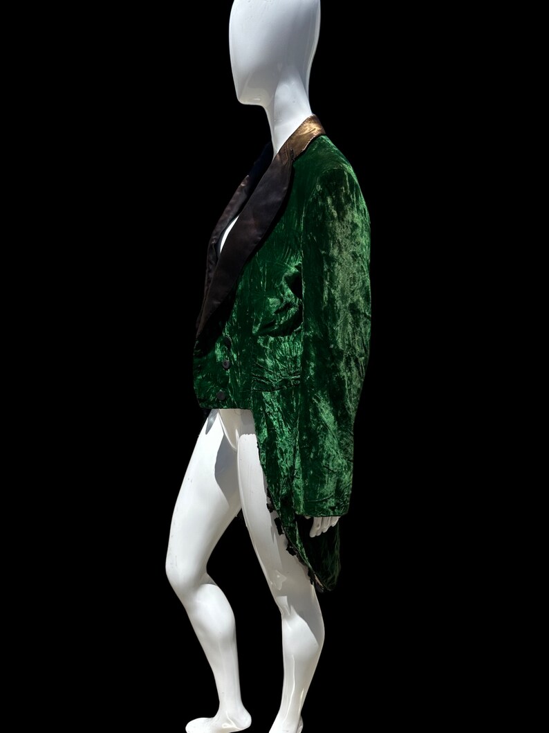 K&ouml;nnte beinhalten: Eine gr&uuml;ne Samtjacke mit schwarzem Satinrevers und goldenen Verzierungen. Die Jacke hat einen langen Schwanz und wird auf einem wei&szlig;en Mannequin pr&auml;sentiert.