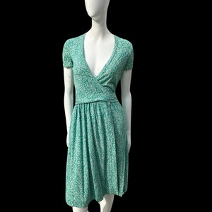 May include: A mint green dress with a white floral pattern. The dress features a V-neckline, short sleeves, and a gathered waist. The skirt falls to the knees. The dress is displayed on a white mannequin against a black background.
