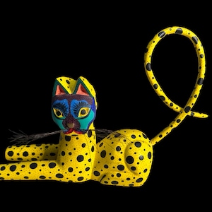 May include: A yellow wooden cat figurine with black spots and a blue, green, and purple painted face. The cat is lying down with its tail curled up in the air.