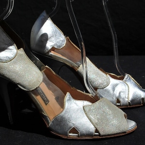 May include: A pair of silver high-heeled shoes with open toes and a sparkly design. The shoes have a delicate strap across the foot and a cut-out design on the side.