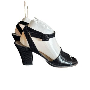 May include: Black open-toe heels with an ankle strap and buckle closure. The heels have a textured pattern and a chunky heel design. The shoes are displayed on a white background.
