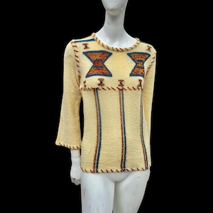 Puede incluir: Un suéter vintage de color crema con cuello redondo y mangas tres cuartos. El suéter presenta un diseño geométrico en tonos azules y naranjas en el pecho, con rayas verticales y costuras decorativas.